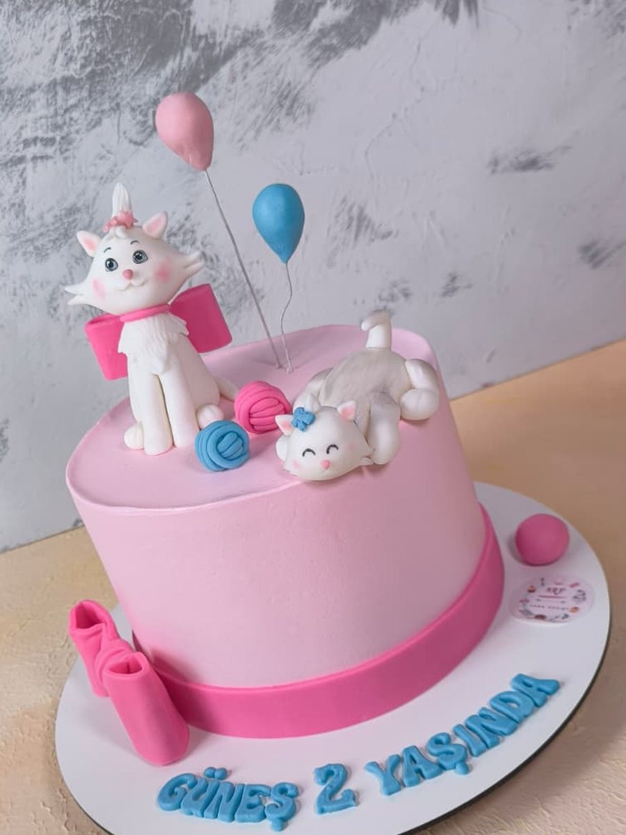 Pembe kremalı, fuşya kurdeleli, üzerinde yün yumaklarıyla oynayan iki beyaz kedi figürü ve balonlar olan doğum günü pastası